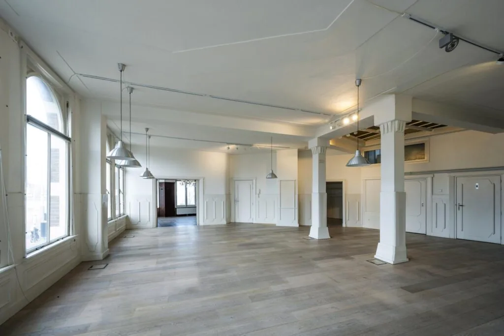 Lichte, leegstaande ruimte aan de De Ruijterkade met houten vloer, witte muren en grote ramen.